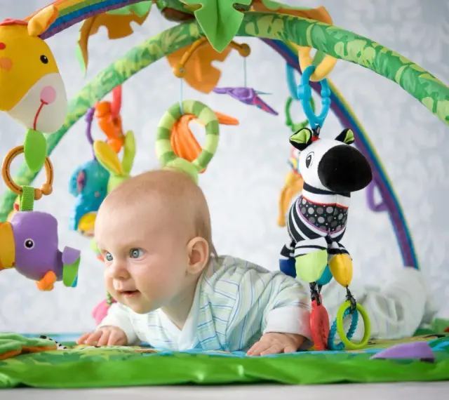Niemowlę leży na kolorowej macie edukacyjnej, bawiąc się wiszącymi zabawkami: żyrafą, papugą i zebrą.