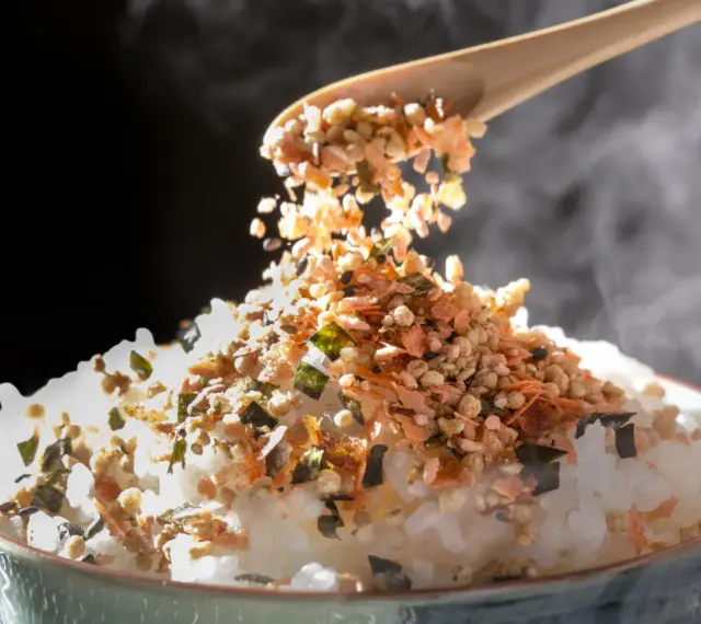 Łyżka sypie na parujący ryż kolorowy furikake. To japońska posypka, która dodaje smaku.