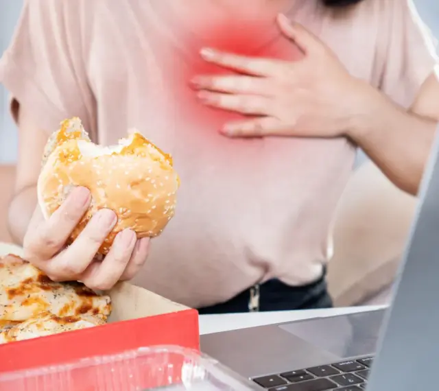 Kobieta z bólem w klatce piersiowej trzyma hamburgera. Zastanawia się, co pić na zgagę po zjedzeniu pizzy i fast foodów.