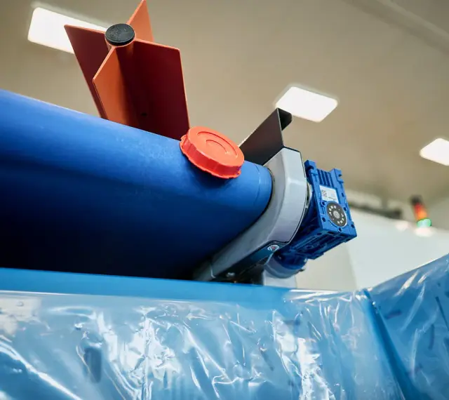 Maszyna do produkcji folii, z niebieskim wałkiem i pomarańczową nakrętką, przygotowuje materiał cerplast do dalszej obróbki.