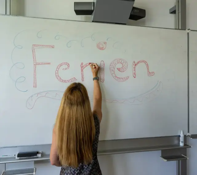 Frau schreibt "Ferien" an Tafel, Vorfreude auf Pfingstferien Rheinland-Pfalz.