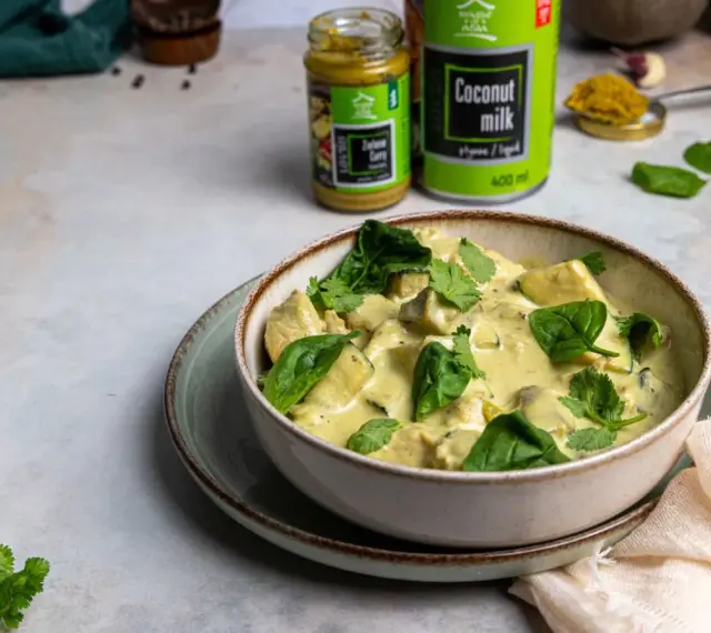 Aksamitny kurczak w zielonym curry, udekorowany świeżym szpinakiem i kolendrą. Obok puszka mleka kokosowego i słoiczek pasty curry.