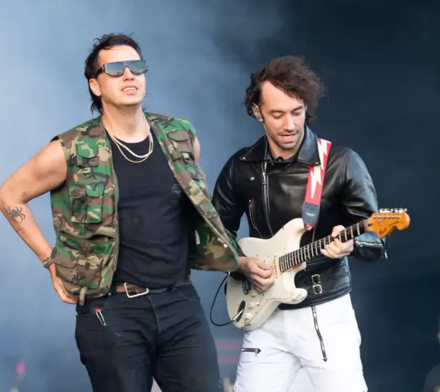 Albert Hammond Jr. w kamizelce moro i okularach stoi obok gitarzysty w skórzanej kurtce, grającego na białej gitarze.