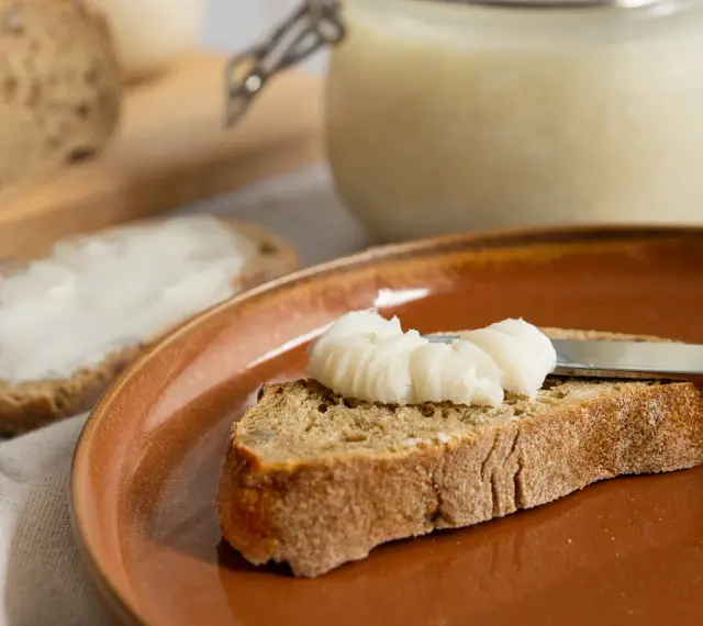 Ein Glas mit selbstgemachtem Schmalz steht bereit. Eine Scheibe Brot wird gerade mit dem Schmalz bestrichen, ein Genuss für jeden, der Schmalz selber machen möchte.