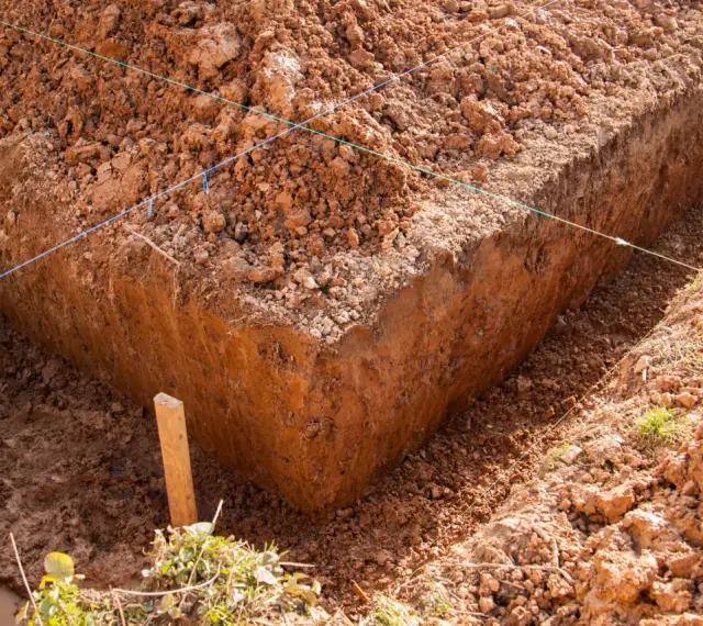 Głęboki wykop pod fundament, z widocznymi nićmi wyznaczającymi linie budowy. Widać błotniste dno i usypaną ziemię.