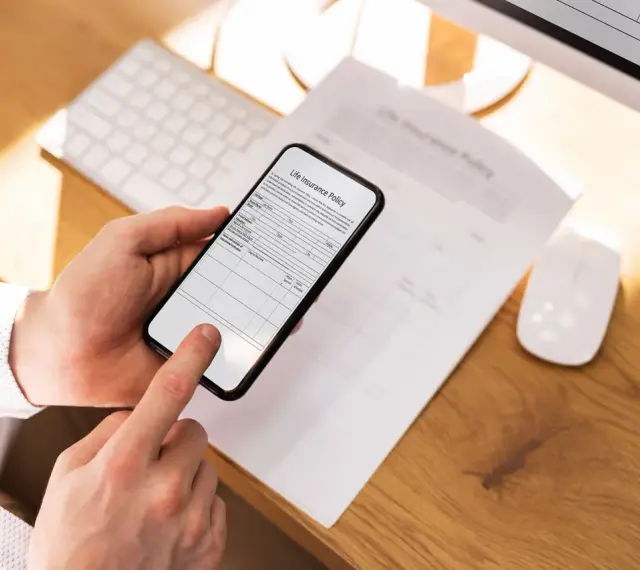 Ręce trzymają smartfon, który działa jak skaner dokumentów, skanując polisę ubezpieczeniową leżącą na biurku.