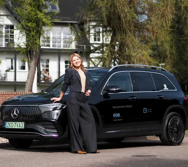 Kobieta w czarnym garniturze opiera się o czarnego Mercedesa EQB na tle białego domu.