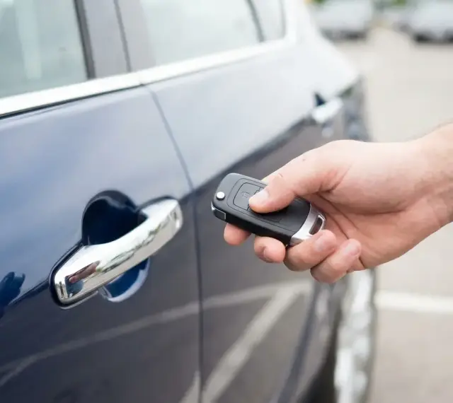 Dłoń trzyma kluczyk keyless, zbliżając go do klamki ciemnoniebieskiego samochodu.