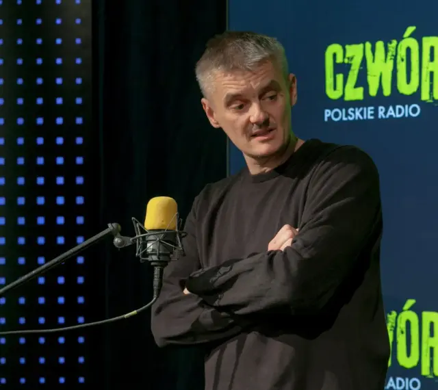 Jacek Sienkiewicz w studiu Polskiego Radia, przed mikrofonem, z rękami skrzyżowanymi na piersi.