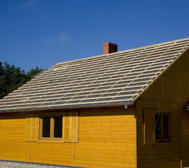 Budowa domu z żółtej deski. Na dachu układana jest papa, przygotowana pod pokrycie.