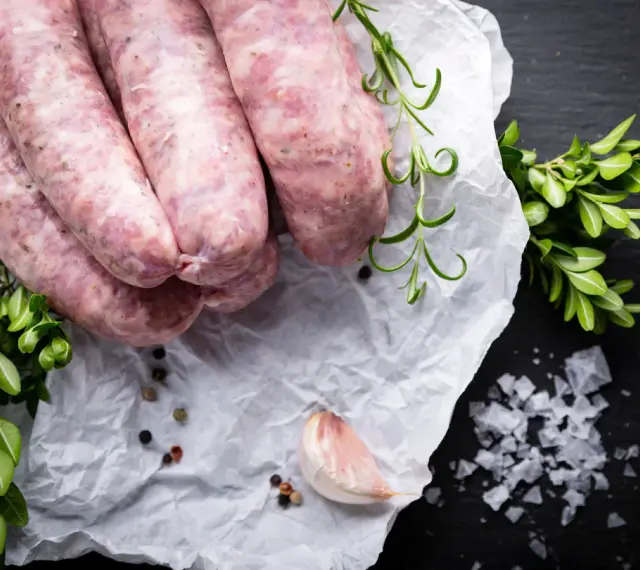 Surowa biała kiełbasa na papierze, obok gałązki rozmarynu i czosnek. Zastanawiasz się nad biała kiełbasa kcal?