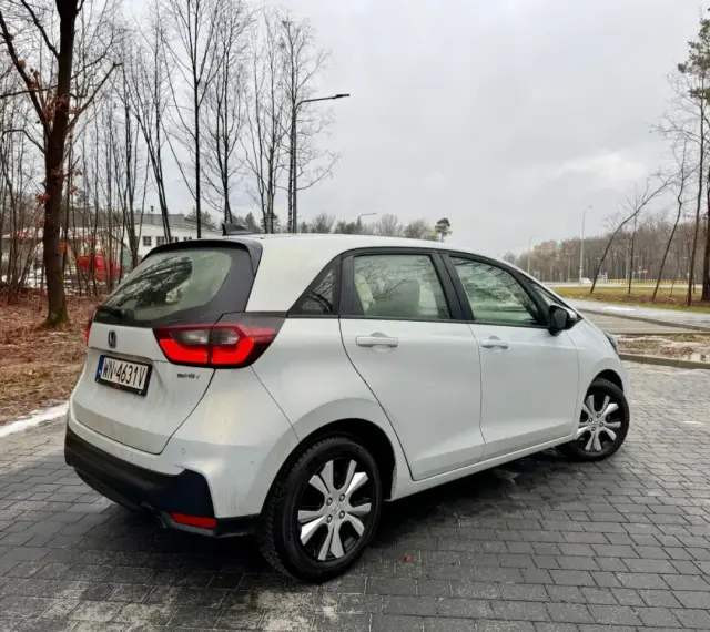 Biały Honda Fit stoi na mokrej, brukowanej nawierzchni. W tle drzewa i budynki.