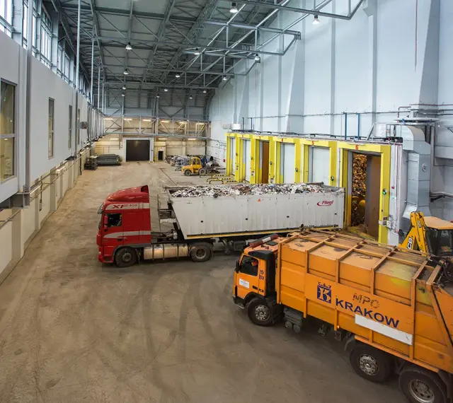 Wielka hala przemysłowa, gdzie trwa termiczne przekształcanie odpadów. Ciężarówki z odpadami czekają na rozładunek, a koparka przygotowuje materiał do przetworzenia.