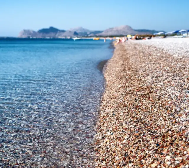 Rodos: turkusowe morze obmywa kamienistą plażę, w tle góry i plażowicze. Idealne miejsce na wakacje.