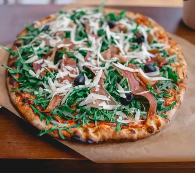 Pyszna pizza z rukolą, oliwkami, parmezanem i cienkimi plastrami prosciutto.