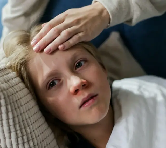 Dziecko z gorączką leży na poduszce, a ręka mamy sprawdza mu czoło. Czy dziecko z gorączką może wychodzić na dwór?