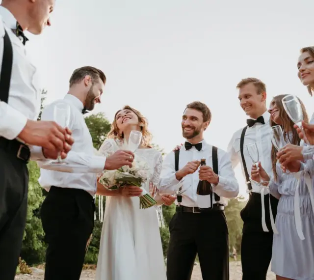 Świadek na ślubie otwiera szampana, podczas gdy panna młoda i jej drużba trzymają kieliszki.