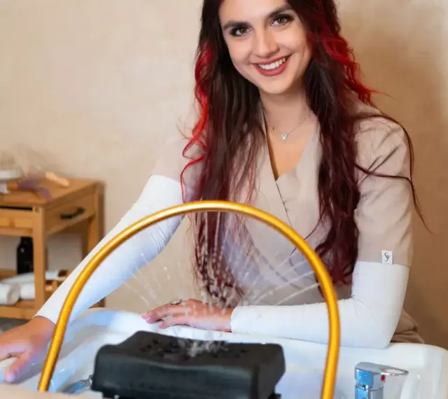Uśmiechnięta stylistka z rudymi pasemkami przygotowuje się do zabiegu. Dowiedz się, ile kosztuje pedicure w tym salonie.