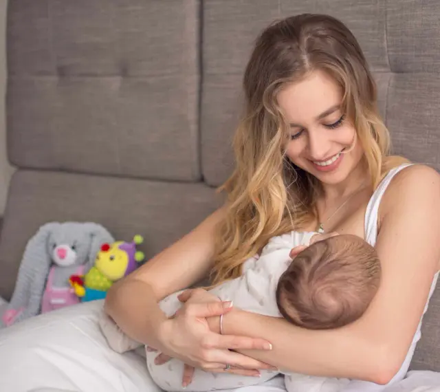 Uśmiechnięta mama karmi piersią dziecko. W tle pluszowe zabawki. Nakładki na pierś do karmienia mogą pomóc w tym intymnym momencie.