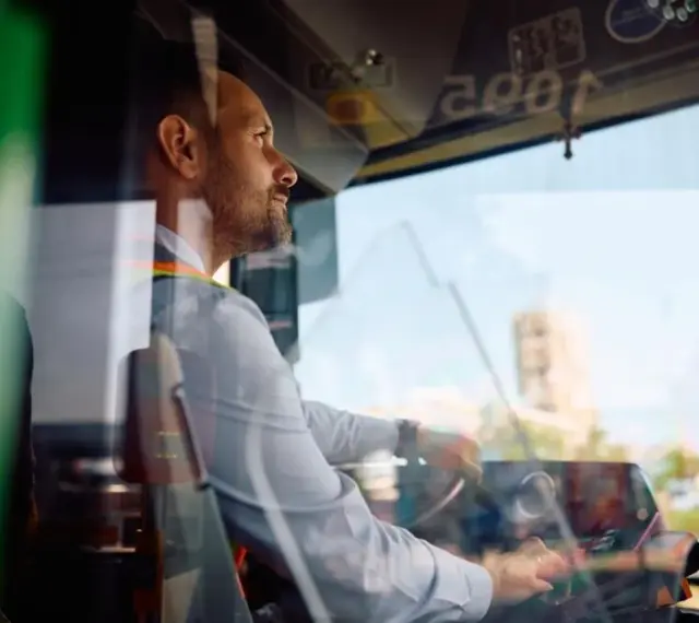 Skupiony kierowca autobusu prowadzi pojazd, trzymając kierownicę. Widać jego profil i fragment deski rozdzielczej.