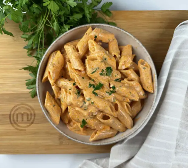 Penne alla vodka cremose e invitanti, guarnite con prezzemolo fresco, servite in una ciotola su un tagliere di legno con il logo MasterChef.
