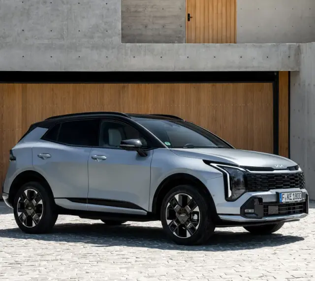 Elegancki, srebrny Kia Sportage Hybrid na tle nowoczesnej architektury z drewnianymi drzwiami i betonowymi schodami.
