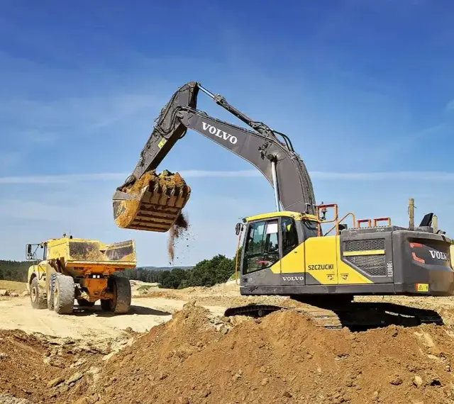 Koparka Volvo wysypuje ziemię do wywrotki. Prace ziemne, ułożenie rur w wykopie cennik bez materiału.