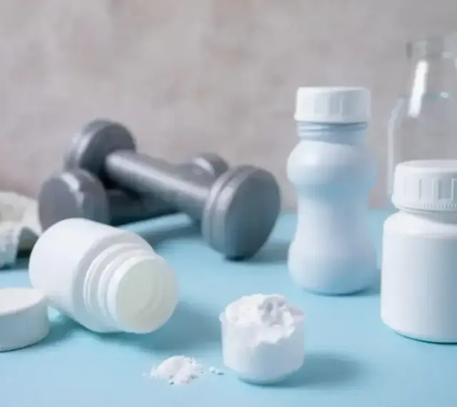 Białe pojemniki, miarka z proszkiem i hantle sugerują suplementację. Proteiny co to? To budulec mięśni, ważny dla sportowców.