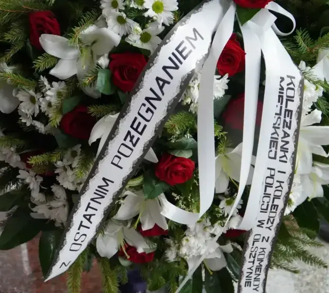 Wieniec pogrzebowy z białymi liliami i czerwonymi różami. Napis na szarfie: "Z ostatnim pożegnaniem od koleżanki i kolegów z Komendy Miejskiej Policji w Olsztynie".