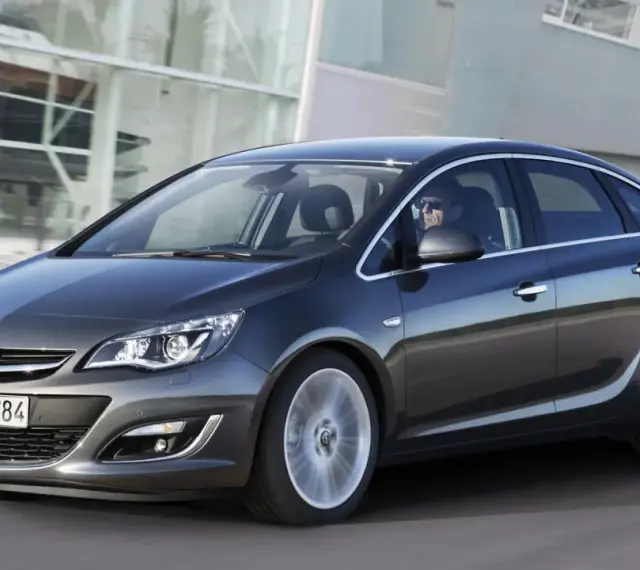 Ciemnoszary Opel Astra J 1.6 benzyna w ruchu, z kierowcą w okularach za kierownicą, na tle nowoczesnego budynku.