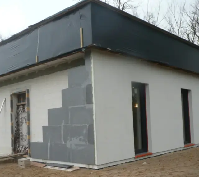 Budowa domu ze styropianu w trakcie. Widoczne bloczki, okna i dach przykryty czarną folią.