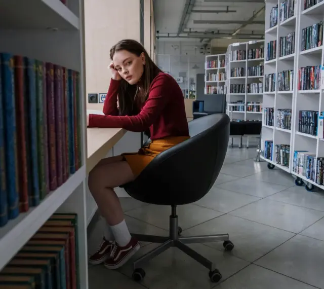 Zamyślona nastolatka w bibliotece, otoczona książkami, odzwierciedla złożoność zachowań nastolatków w okresie dojrzewania.