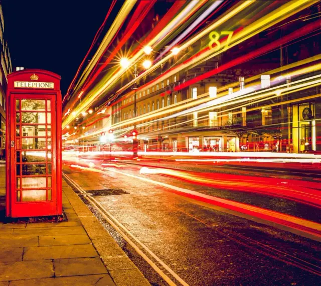 Klasyczna czerwona budka telefoniczna na tle nocnej ulicy, gdzie długi czas naswietlania zdjecia uchwycił smugi świateł samochodowych.