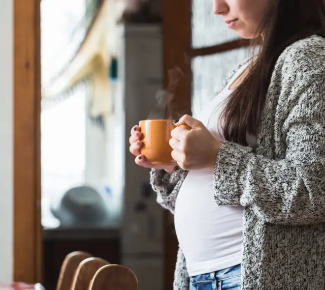 Ciepły napar z rooibos w dłoniach ciężarnej kobiety. Delikatny dym unosi się z kubka, tworząc chwilę spokoju.