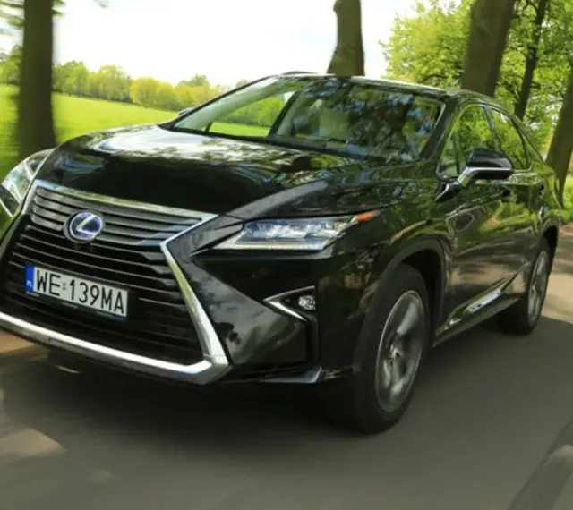 Czarny Lexus RX 450h pędzi po wiejskiej drodze, otoczony zielenią drzew i pól.