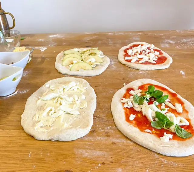 Vier vorbereitete Pinsa-Teiglinge liegen auf einem Holztisch. Einer mit Apfelscheiben, einer mit Tomatensoße und Mozzarella, einer nur mit Mozzarella und einer mit Tomatensoße, Mozzarella und Basilikum.