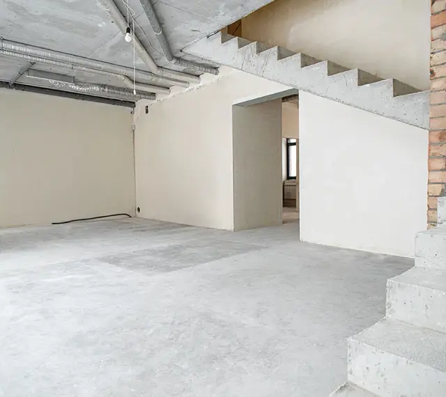 Wnętrze w stanie deweloperskim: betonowe schody, nieotynkowane ściany, widoczne instalacje i przewody.