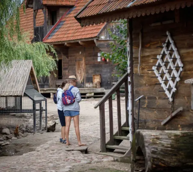 Dwie dziewczyny zwiedzają skansen, który może być częścią oferty hotelu bon turystyczny. W tle drewniane chaty i kurnik.