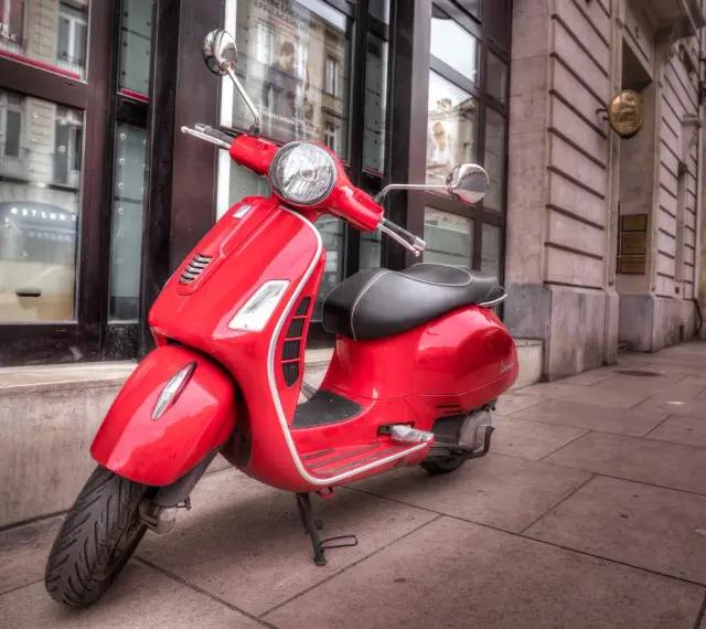 Czerwony skuter Vespa, idealny do zdobycia prawa jazdy AM, stoi na chodniku przed budynkiem.