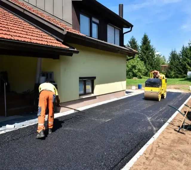 Pracownik równa łopatą świeży asfalt na podjazd, a walec go ubija. Dom w tle.