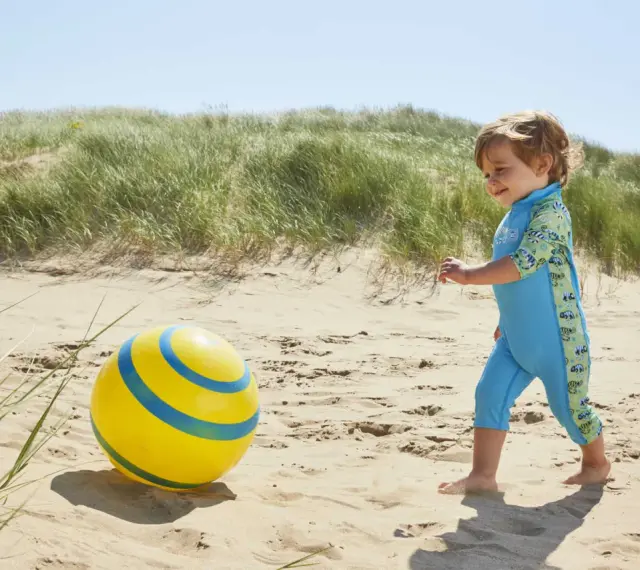 Chłopiec w niebieskim stroju kąpielowym z neoprenu bawi się żółtą piłką na plaży.