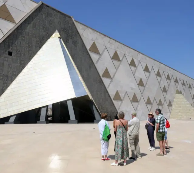 Grupa turystów podziwia nowoczesne muzeum w Gizie, którego fasada zdobiona jest hieroglifami i geometrycznymi wzorami przypominającymi piramidy.