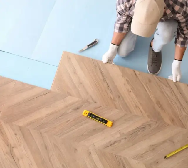 Instalator układa panele podłogowe w jodełkę. Widoczny poziomica i miarka.