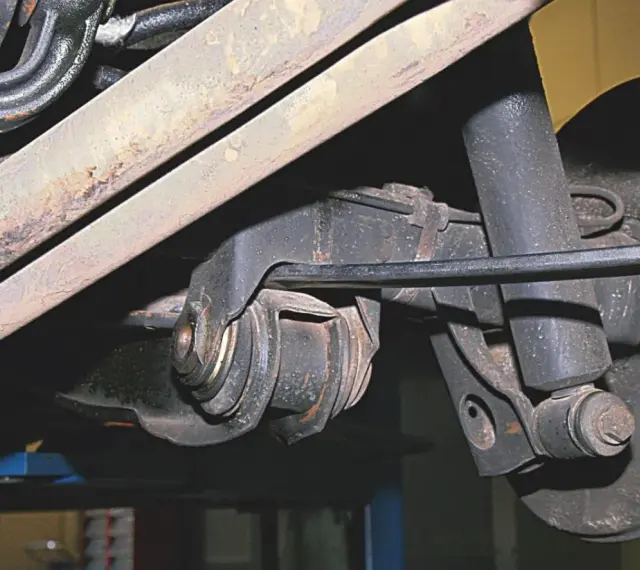Widok z dołu na element zawieszenia samochodu. Widoczny jest metalowy wahacz z gumowymi tulejami, amortyzator i fragment układu wydechowego.