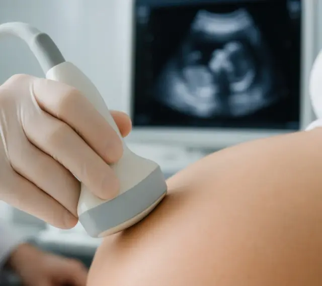 Ręka w rękawiczce medycznej wykonuje usg prenatalne. Na ekranie widoczny obraz płodu.