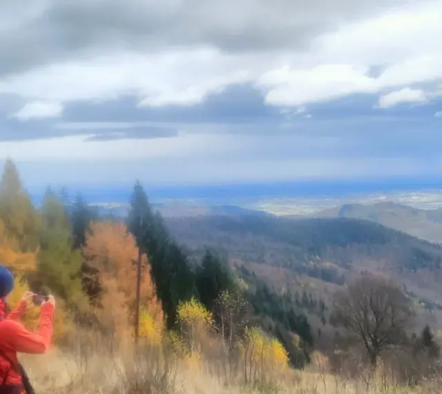 Widok na Beskid Mały jesienią. Turysta z pomarańczowym plecakiem fotografuje panoramę.