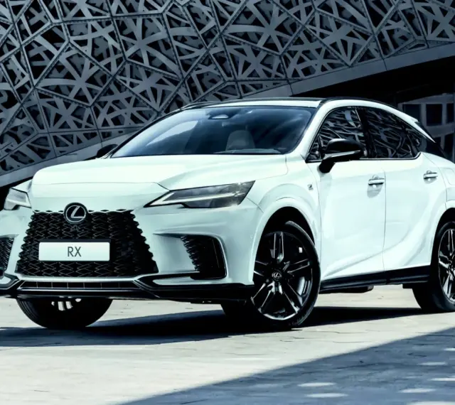 Biały Lexus RX500h z czarnymi felgami stoi przed nowoczesnym budynkiem o geometrycznej fasadzie.