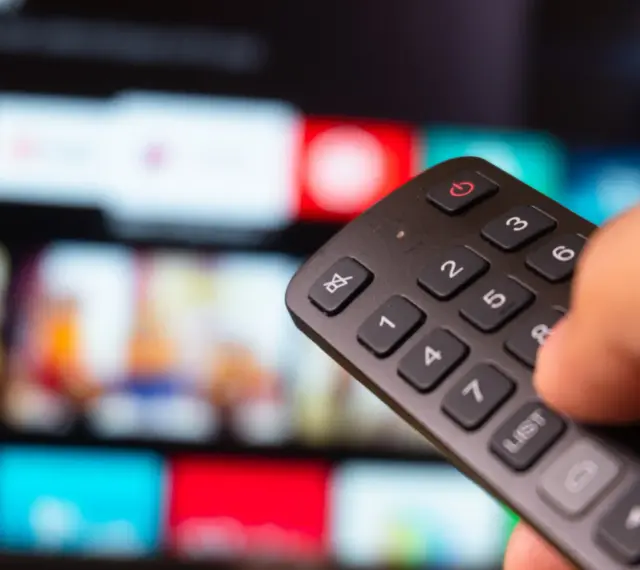 Dłoń trzyma pilot do telewizora, wybierając film na ekranie. To idealny moment na relaks z web tv.
