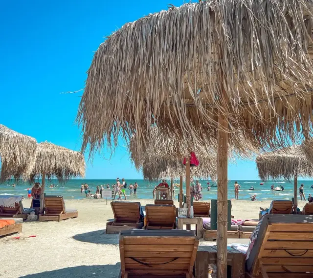 Słoneczne rumuńskie wakacje nad morzem. Leżaki i parasole z trawy na piaszczystej plaży, w tle ludzie kąpiący się w morzu.