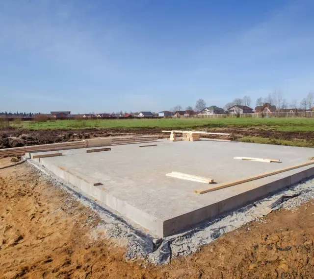 Płyta fundamentowa, jeden z rodzajów fundamentów, gotowa pod budowę domu. W tle domy i błękitne niebo.